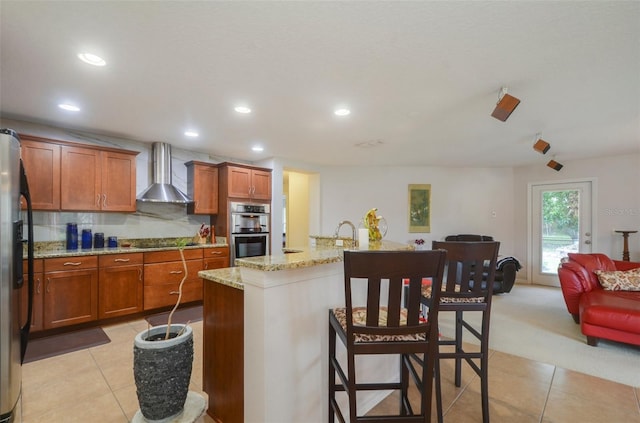 kitchen featuring backsplash, a kitchen island with sink, a breakfast bar area, wall chimney exhaust hood, and stainless steel appliances