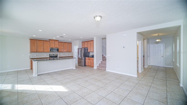 kitchen with appliances with stainless steel finishes, backsplash, a textured ceiling, a kitchen island with sink, and light tile patterned floors