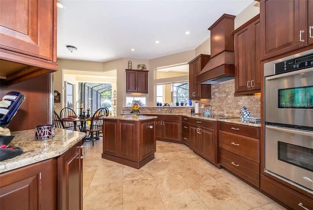 kitchen with light stone countertops, sink, stainless steel double oven, a kitchen island, and custom range hood