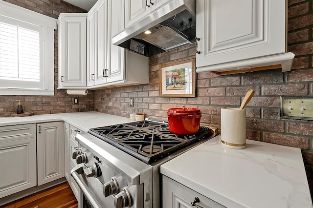 kitchen featuring under cabinet range hood, stainless steel range with gas stovetop, brick wall, and white cabinetry