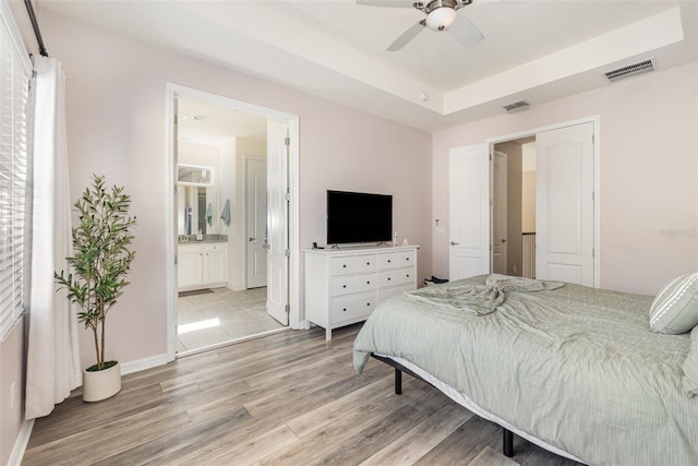 bedroom with ceiling fan, light hardwood / wood-style floors, ensuite bath, and a tray ceiling