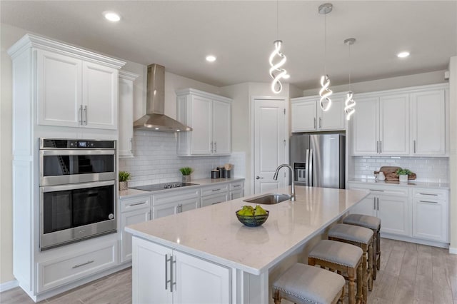 kitchen with a center island with sink, white cabinets, appliances with stainless steel finishes, wall chimney range hood, and a sink