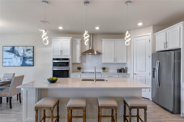 kitchen featuring a kitchen island with sink, white cabinetry, appliances with stainless steel finishes, wall chimney range hood, and pendant lighting