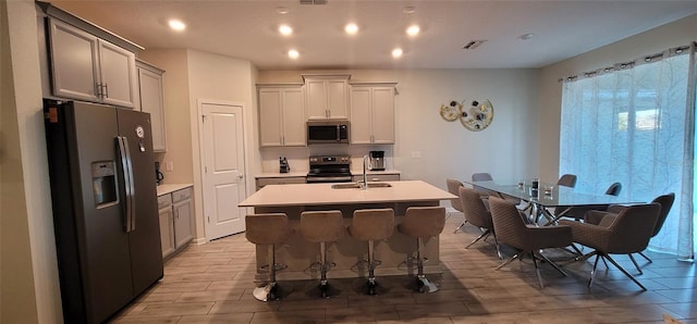 kitchen with sink, gray cabinets, appliances with stainless steel finishes, a kitchen island with sink, and a kitchen breakfast bar