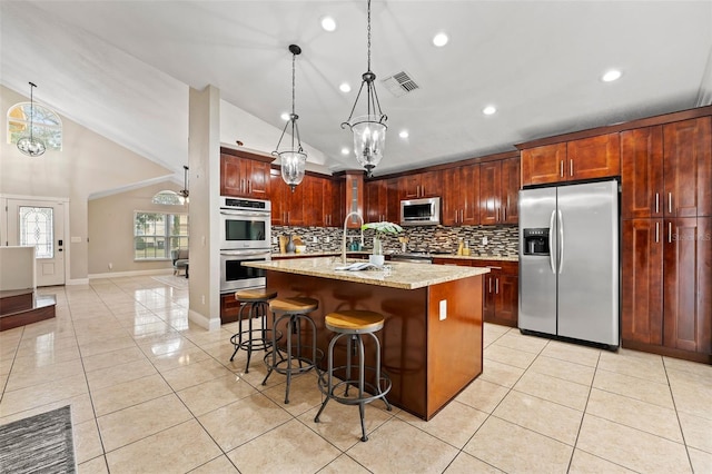 kitchen with pendant lighting, a kitchen bar, an island with sink, appliances with stainless steel finishes, and light stone counters