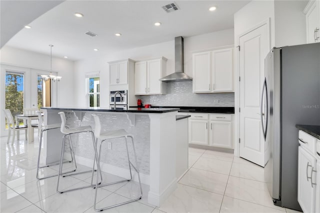 kitchen with stainless steel appliances, a kitchen island with sink, wall chimney range hood, decorative light fixtures, and white cabinetry