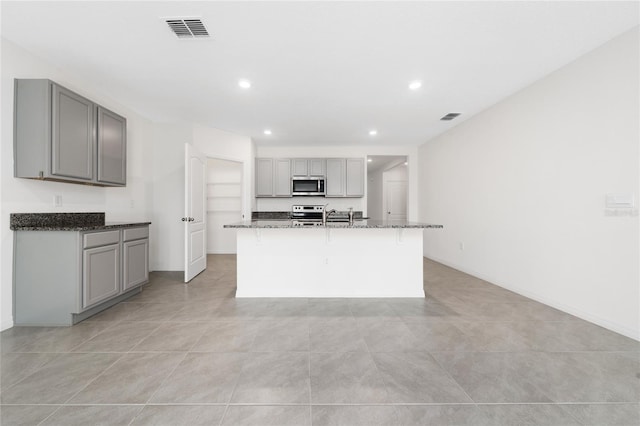 kitchen featuring dark stone counters, stainless steel appliances, gray cabinets, a breakfast bar area, and an island with sink