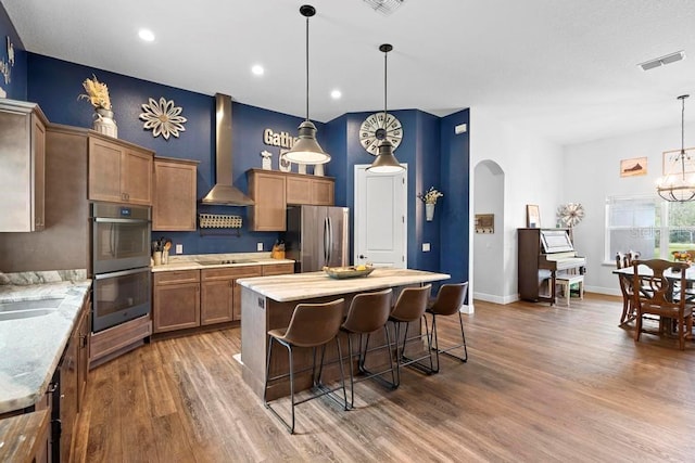 kitchen with hanging light fixtures, appliances with stainless steel finishes, brown cabinetry, and wall chimney range hood