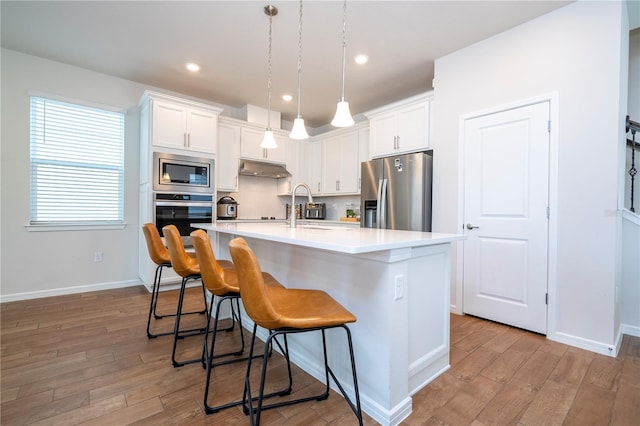 kitchen featuring decorative backsplash, stainless steel appliances, white cabinets, hanging light fixtures, and an island with sink