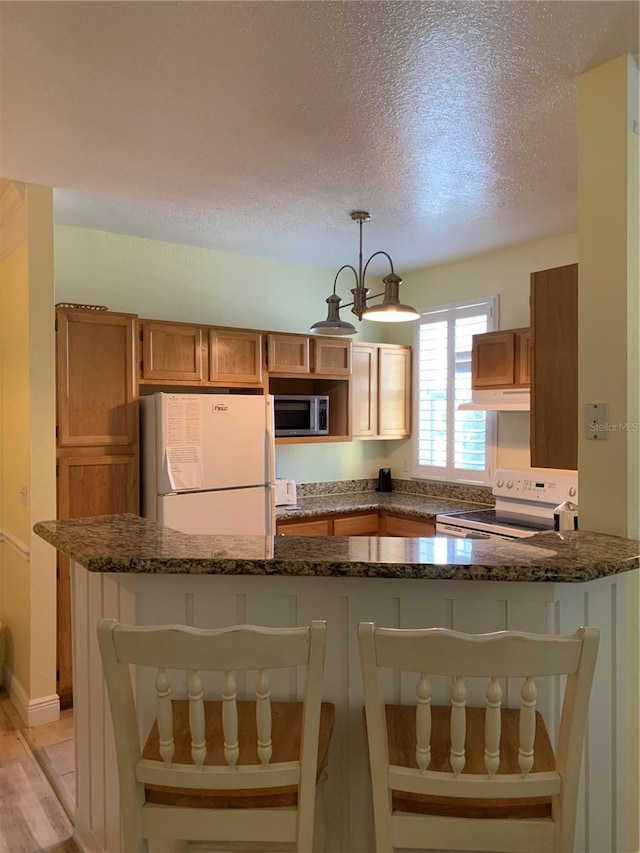 kitchen featuring pendant lighting, brown cabinetry, dark stone countertops, white appliances, and under cabinet range hood