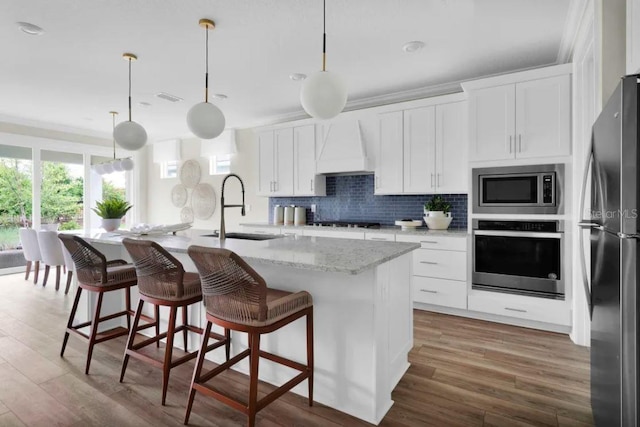 kitchen featuring premium range hood, stainless steel appliances, sink, white cabinetry, and an island with sink