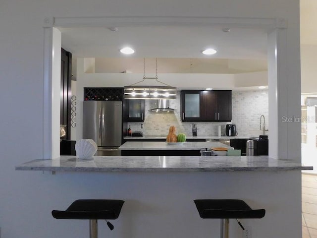 kitchen with dark brown cabinetry, stainless steel fridge, kitchen peninsula, light stone countertops, and decorative backsplash