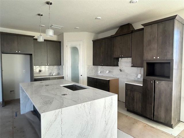 kitchen featuring hanging light fixtures, backsplash, a textured ceiling, a kitchen island with sink, and dark brown cabinets