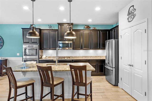 kitchen featuring stainless steel appliances, hanging light fixtures, a center island with sink, and light stone counters