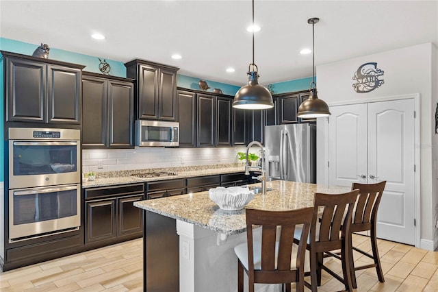 kitchen with pendant lighting, stainless steel appliances, light stone countertops, an island with sink, and decorative backsplash