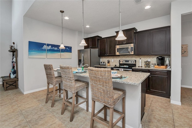 kitchen with a breakfast bar, stainless steel appliances, an island with sink, decorative light fixtures, and dark brown cabinets