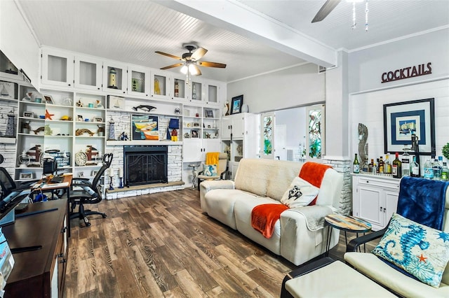 living room with built in shelves, ceiling fan, dark wood-type flooring, a stone fireplace, and ornamental molding