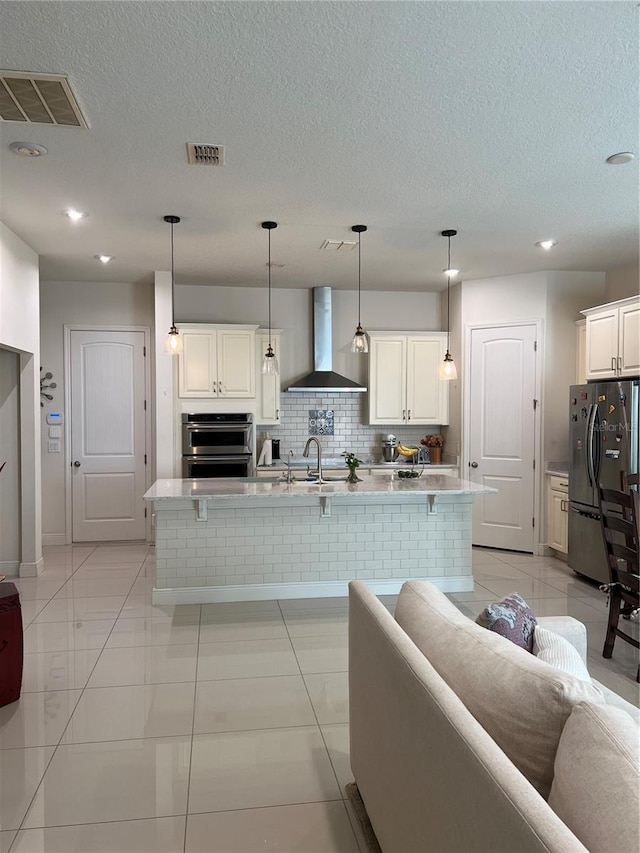 kitchen with a center island with sink, light tile patterned floors, wall chimney range hood, and appliances with stainless steel finishes