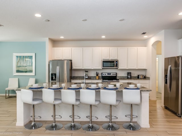 kitchen featuring white cabinetry, stainless steel appliances, a large island with sink, dark stone countertops, and a breakfast bar area