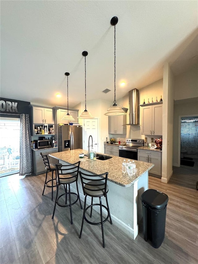 kitchen with gray cabinetry, sink, stainless steel appliances, wall chimney range hood, and a kitchen island with sink