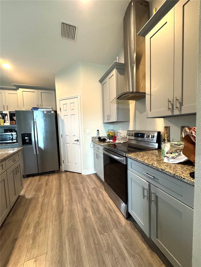 kitchen featuring appliances with stainless steel finishes, dark stone counters, wall chimney exhaust hood, gray cabinetry, and light hardwood / wood-style floors