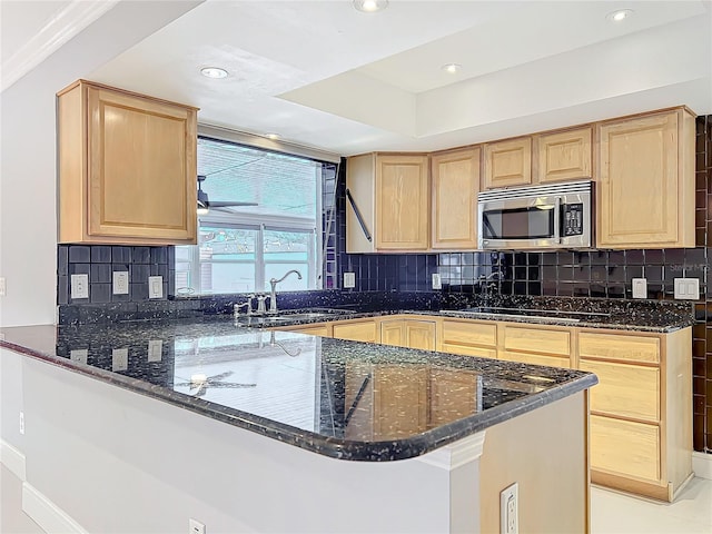 kitchen with stainless steel microwave, backsplash, dark stone countertops, a peninsula, and a sink