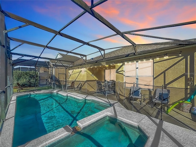 pool at dusk with glass enclosure, a patio area, and an in ground hot tub