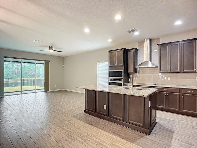 kitchen featuring a kitchen island with sink, ceiling fan, dark brown cabinetry, oven, and wall chimney range hood