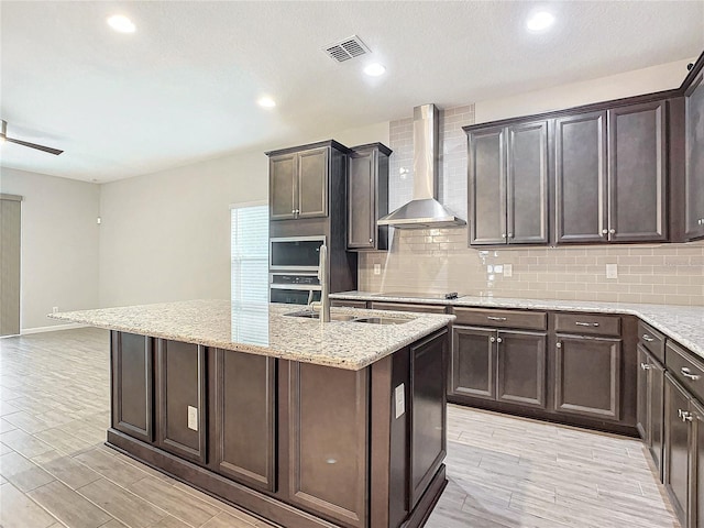 kitchen featuring wall chimney range hood, dark brown cabinetry, an island with sink, light stone countertops, and black electric stovetop