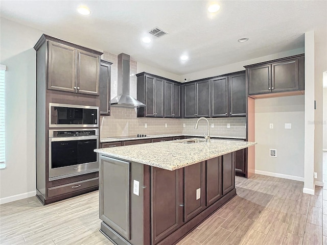 kitchen with a kitchen island with sink, stainless steel appliances, light stone counters, sink, and wall chimney range hood