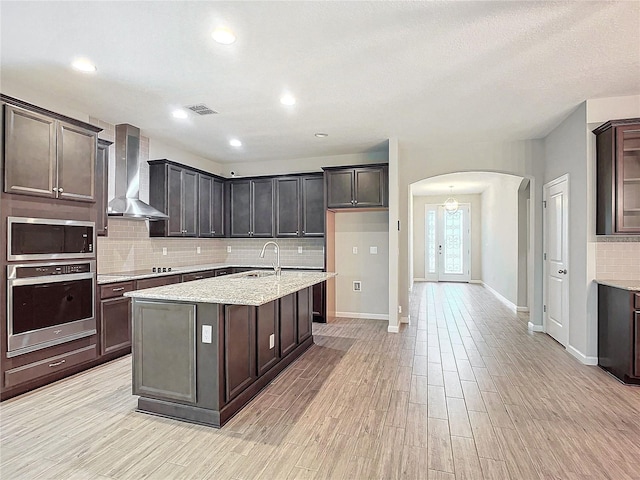 kitchen featuring wall chimney exhaust hood, an island with sink, light wood-type flooring, appliances with stainless steel finishes, and sink