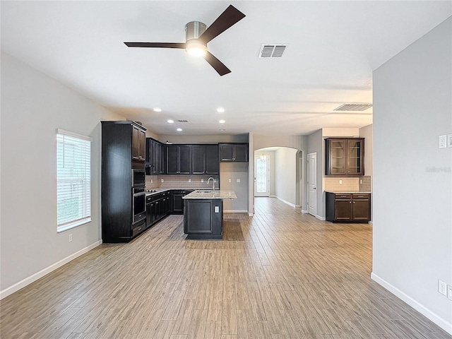 kitchen with sink, ceiling fan, light hardwood / wood-style flooring, backsplash, and a center island with sink