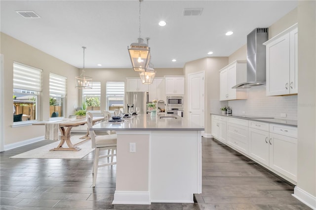 kitchen with appliances with stainless steel finishes, wall chimney exhaust hood, decorative light fixtures, white cabinetry, and an island with sink