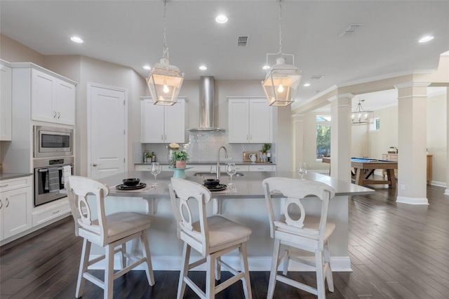 kitchen with white cabinetry, wall chimney range hood, pendant lighting, a center island with sink, and appliances with stainless steel finishes