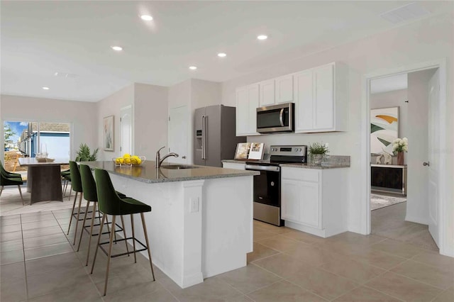 kitchen with a kitchen island with sink, dark stone counters, sink, appliances with stainless steel finishes, and white cabinetry