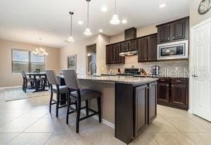 kitchen featuring stainless steel microwave, a kitchen breakfast bar, an island with sink, decorative light fixtures, and light tile patterned floors