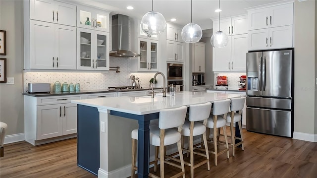 kitchen with white cabinets, wall chimney range hood, a kitchen island with sink, and appliances with stainless steel finishes