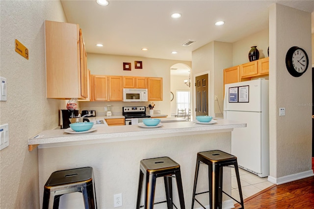 kitchen featuring kitchen peninsula, a kitchen bar, light brown cabinetry, and white appliances