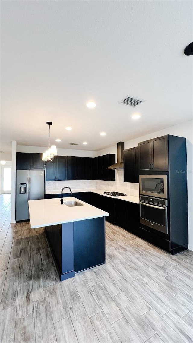 kitchen with wall chimney range hood, stainless steel appliances, sink, hanging light fixtures, and a kitchen island with sink