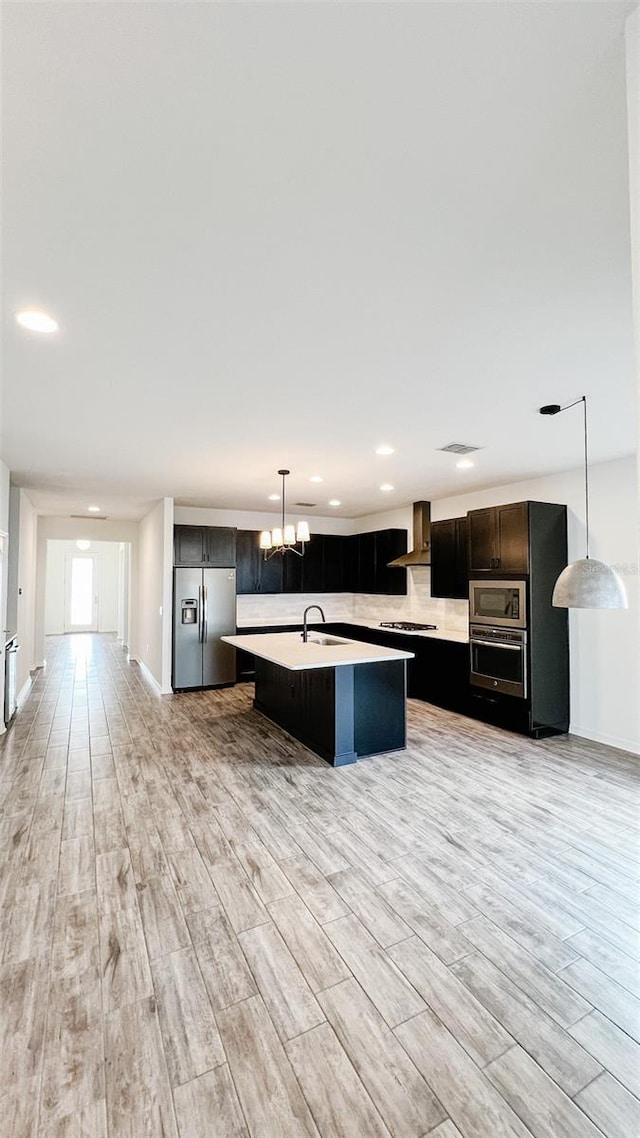 kitchen with pendant lighting, wall chimney range hood, stainless steel appliances, a kitchen island with sink, and a notable chandelier