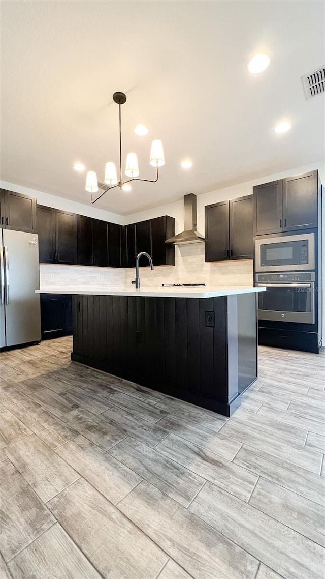 kitchen featuring hanging light fixtures, wall chimney range hood, stainless steel appliances, and a center island with sink