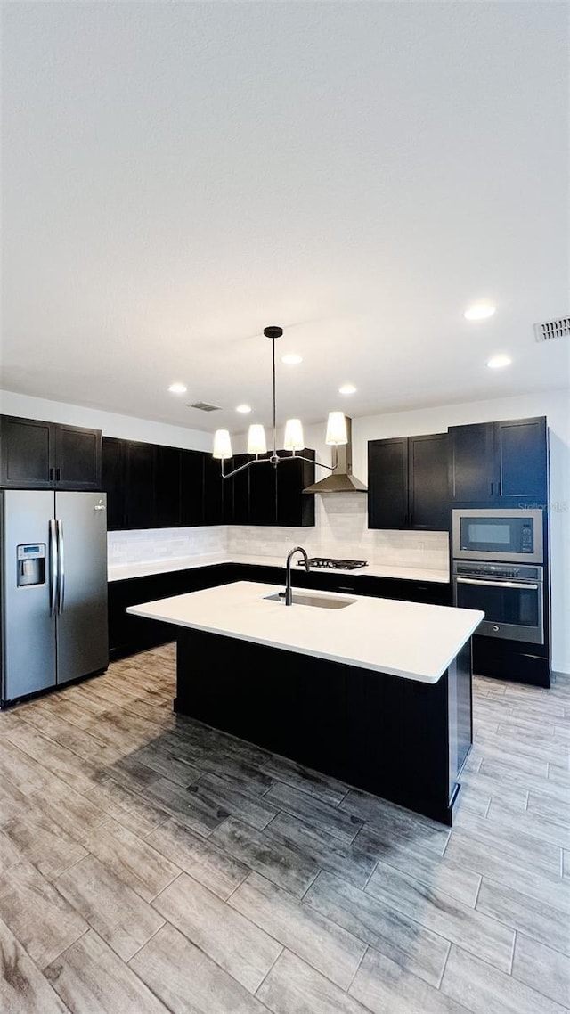 kitchen featuring pendant lighting, wall chimney exhaust hood, stainless steel appliances, an island with sink, and sink