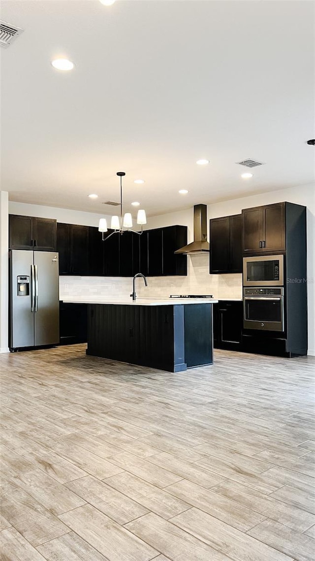 kitchen featuring decorative light fixtures, wall chimney range hood, light hardwood / wood-style floors, a center island with sink, and stainless steel appliances