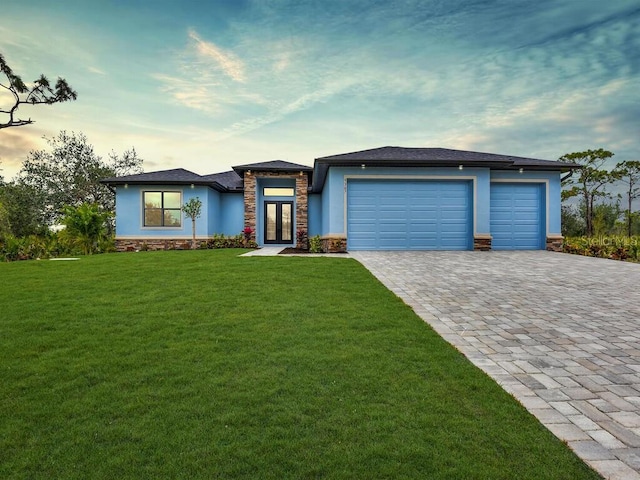 prairie-style home featuring stucco siding, decorative driveway, a yard, stone siding, and an attached garage