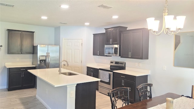 kitchen featuring appliances with stainless steel finishes, a kitchen island with sink, sink, a notable chandelier, and light hardwood / wood-style floors