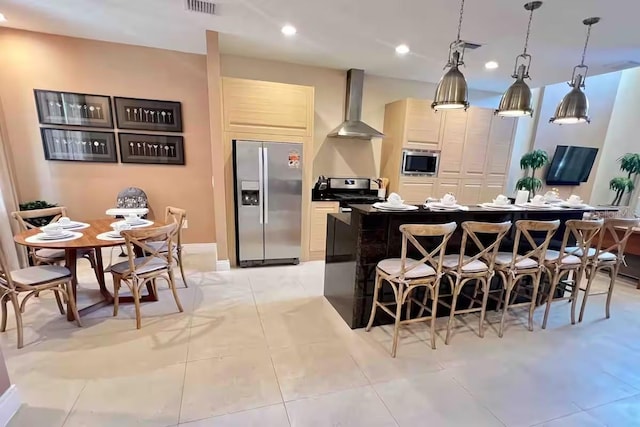 kitchen featuring a center island, wall chimney exhaust hood, a kitchen bar, light tile patterned floors, and appliances with stainless steel finishes