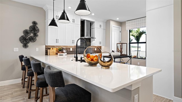 kitchen with kitchen peninsula, hanging light fixtures, wall chimney range hood, white cabinets, and a kitchen breakfast bar