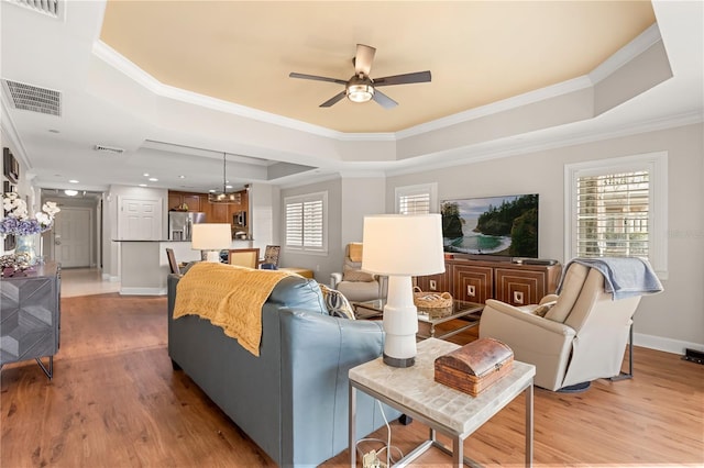 living room with ceiling fan with notable chandelier, ornamental molding, a tray ceiling, and light wood-type flooring