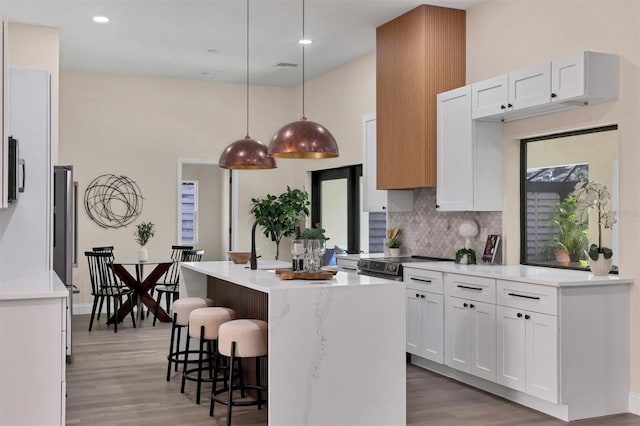 kitchen featuring backsplash, wood-type flooring, decorative light fixtures, a center island with sink, and white cabinetry