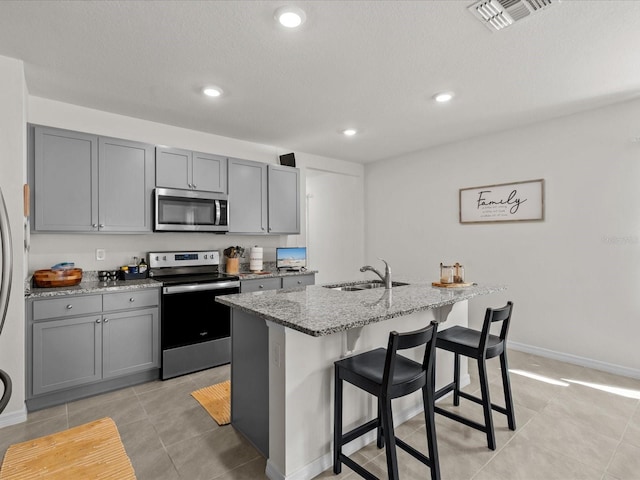 kitchen with sink, gray cabinets, an island with sink, light stone counters, and stainless steel appliances